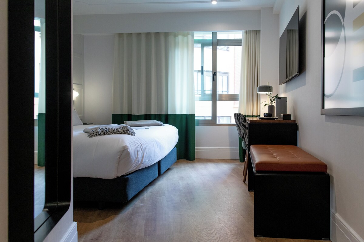 Un dormitorio con cama, un banco, un escritorio, un televisor y una ventana con cortinas.