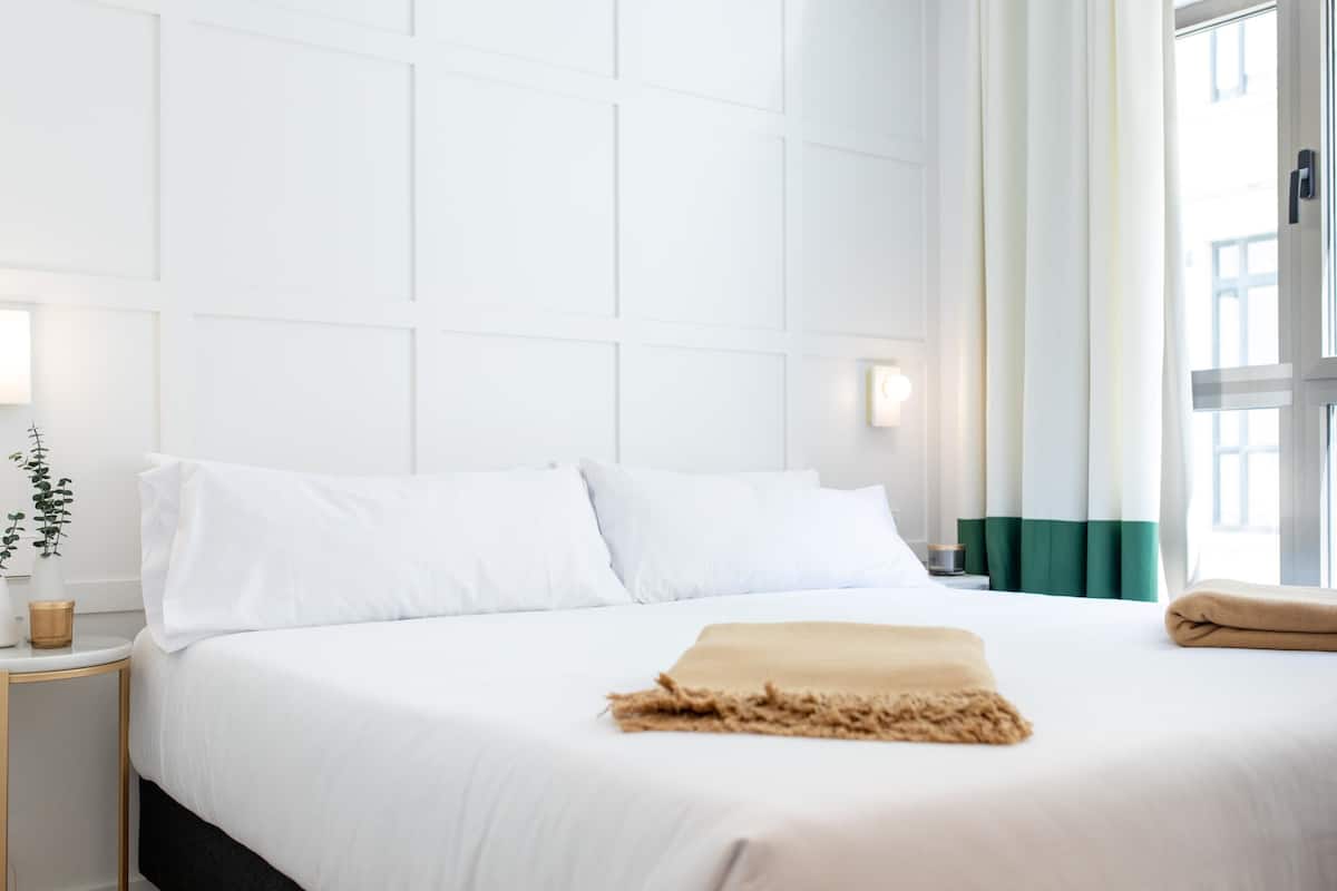 Una cama bien hecha con sábanas blancas, una manta color beige y una cortina verde entreabierta.