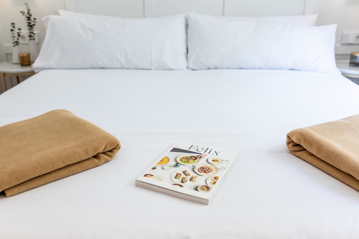 Una cama bien tendida con ropa de cama blanca y dos toallas marrones colocadas a ambos lados de un libro titulado “Félix” con imágenes de comida.