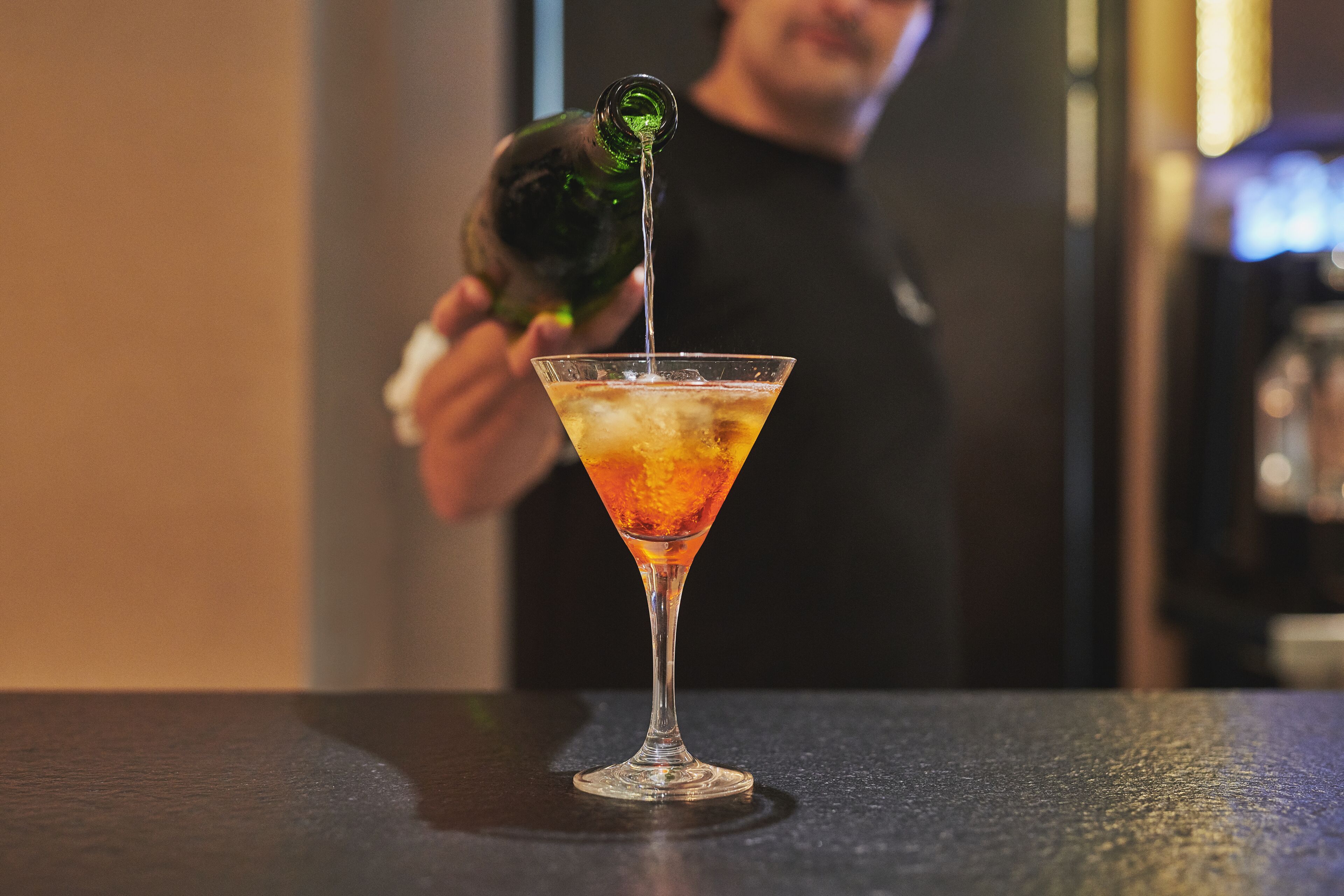 Un verre à martini posé sur le comptoir d’un bar, et une personne qui y verse une boisson.