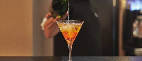 A martini glass on a bar counter with a person pouring a drink into it.
