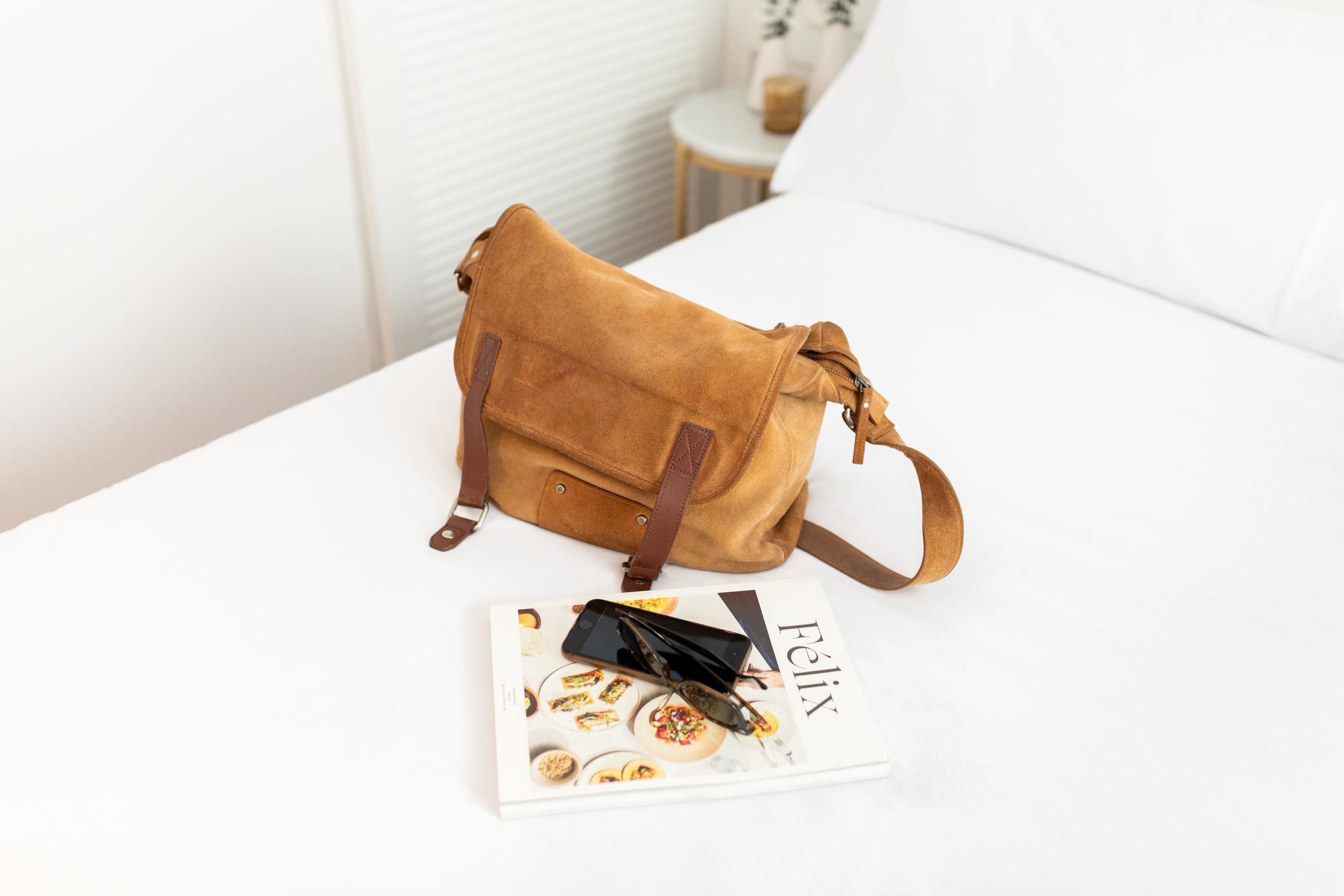 Un sac en daim marron sur un lit blanc, avec un magazine et des lunettes de soleil.