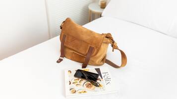 Un sac en daim marron sur un lit blanc, avec un magazine et des lunettes de soleil.
