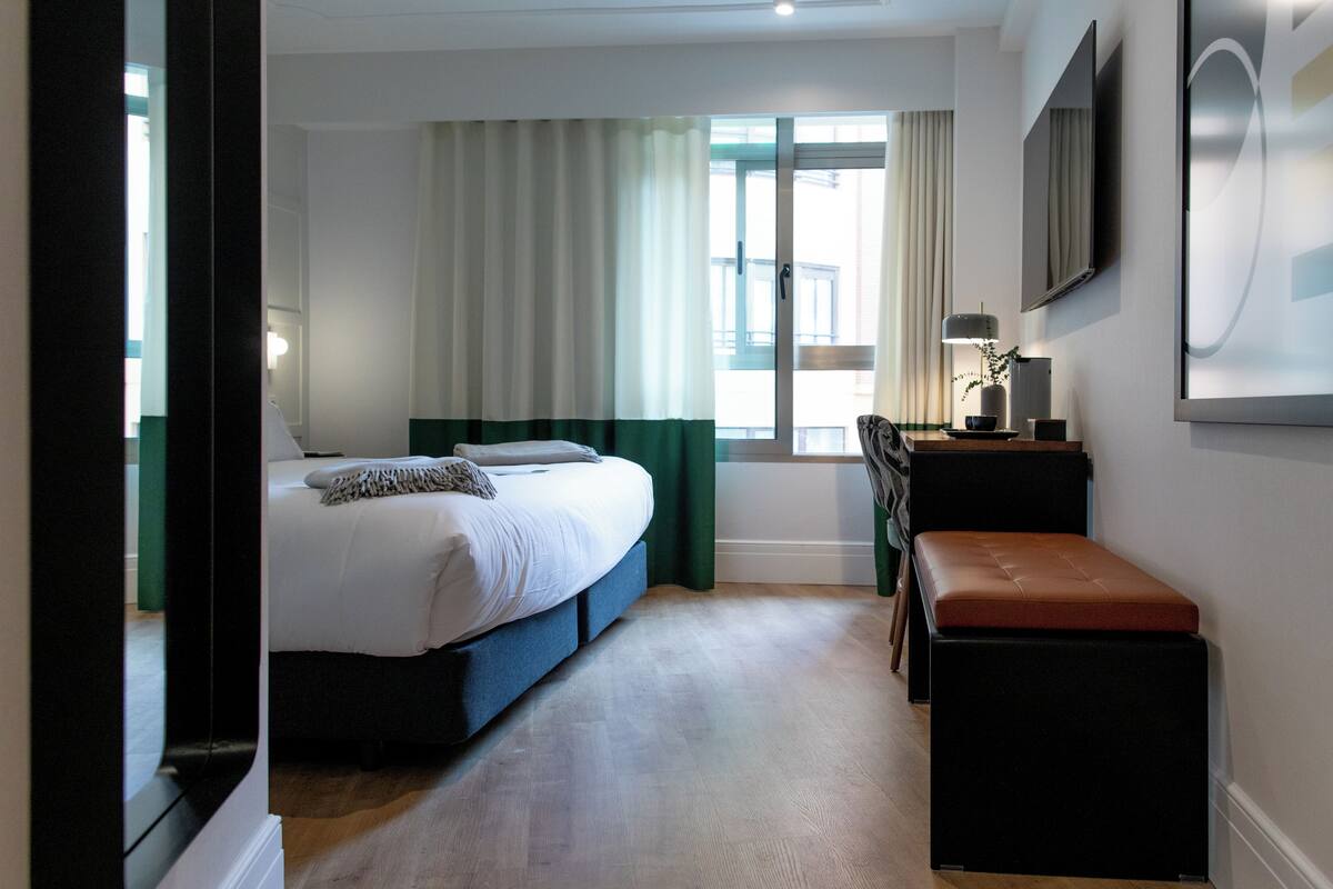 Un dormitorio con cama, un banco, un escritorio, un televisor y una ventana con cortinas.