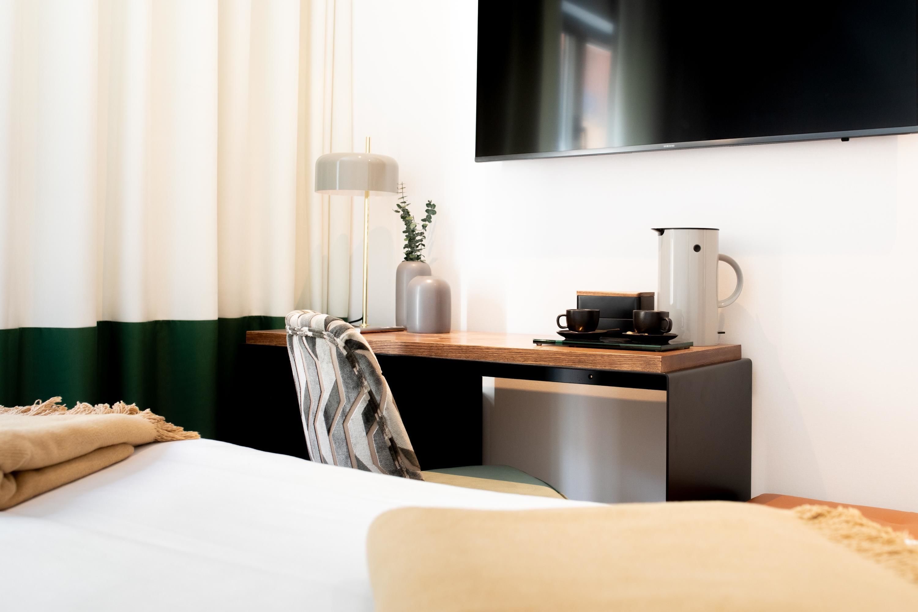 A hotel room with a bed, a desk with a coffee maker, a TV mounted on the wall, and a window with curtains.