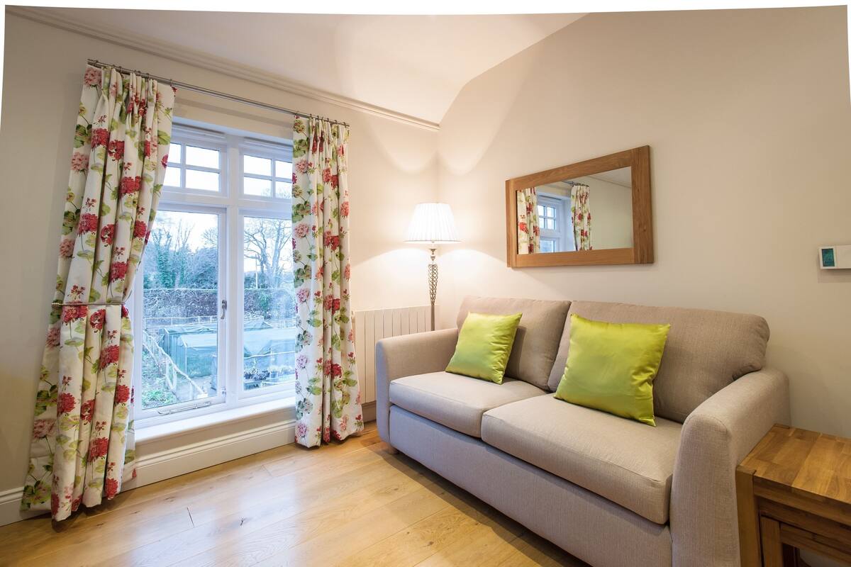 Une pièce avec un canapé beige, des coussins verts, une table d’appoint en bois, un lampadaire et une fenêtre ornée de rideaux à motifs floraux.
