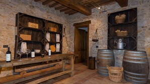 Une cave rustique avec des étagères en bois où sont exposées des bouteilles de vin et des verres, des murs en pierre et de grands tonneaux.