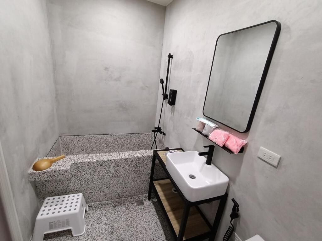 A compact bathroom with a white sink, a mirror, a shower area, and a small shelf with a pink towel.