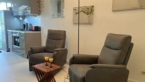 A modern living room with two reclining chairs, a small wooden table with two glasses of orange juice, and a kitchen in the background. - Beach 100m away ▪ Sea view ▪ Apartment classified 3*quiet▪ garden with trees ▪1 bedroom (Le Moule)