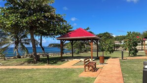 A park with a wooden bench, a red-roofed gazebo, and a pathway leading to the beach. - Beach 100m away ▪ Sea view ▪ Apartment classified 3*quiet▪ garden with trees ▪1 bedroom (Le Moule)
