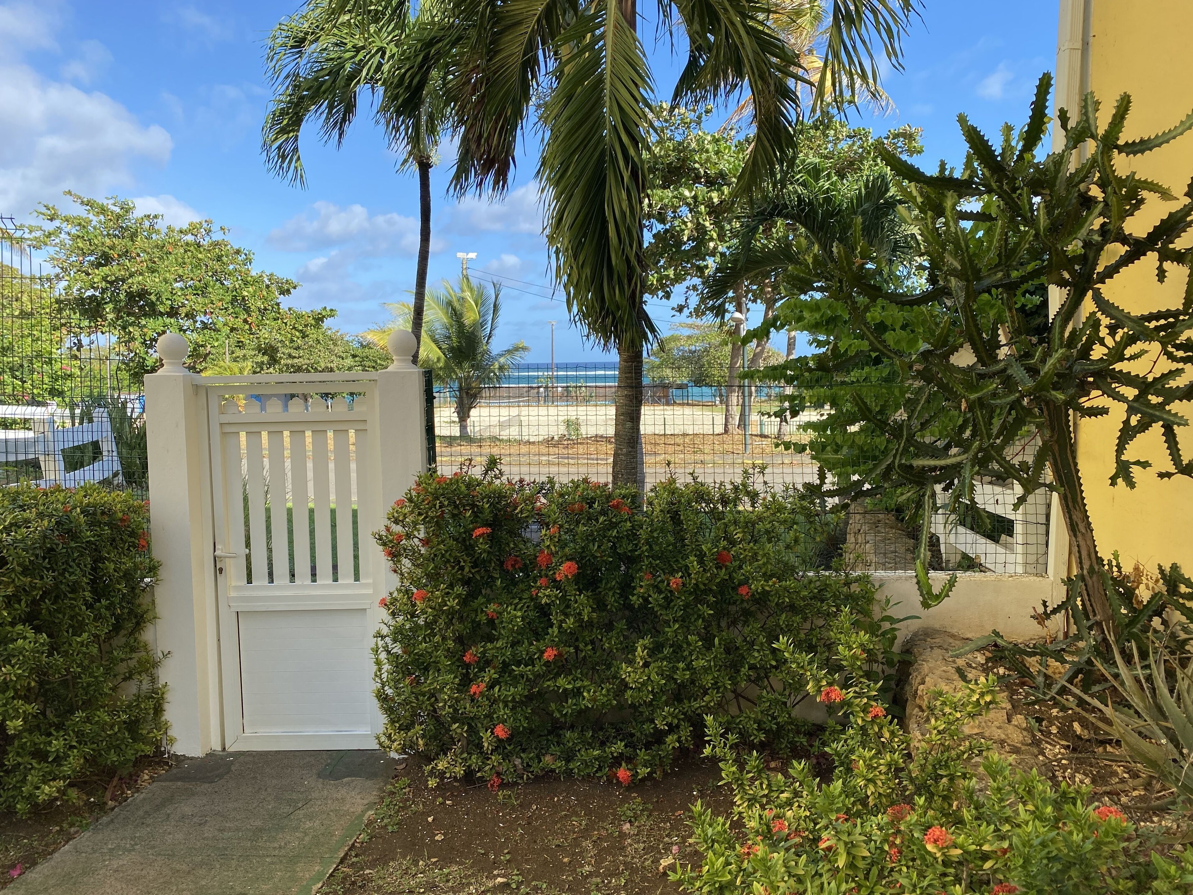 A gated entrance with a white gate, lush greenery, and a view of the beach and ocean.