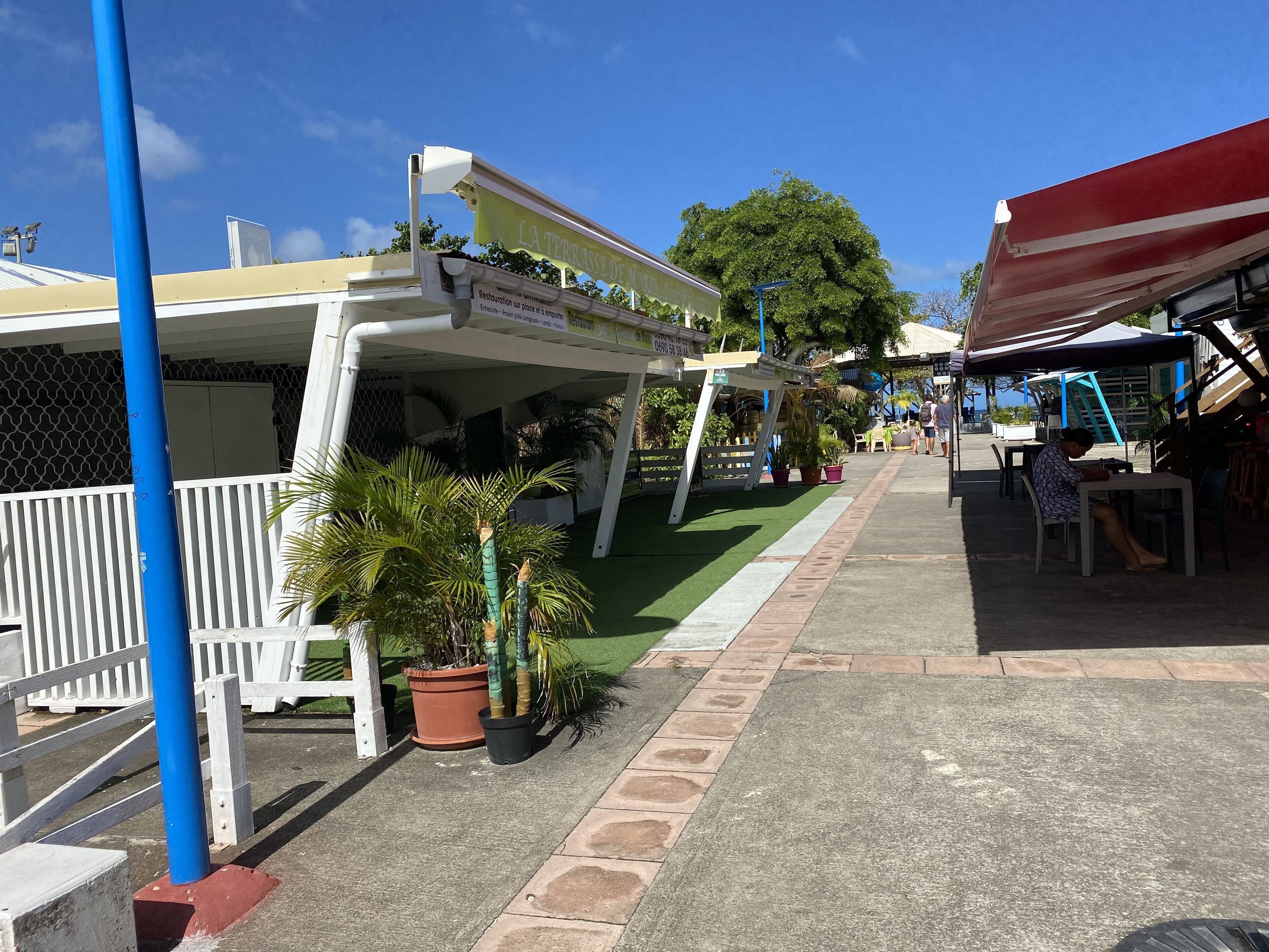 A covered outdoor seating area with tables and chairs, surrounded by potted plants and a white fence.