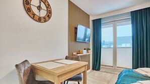 A hotel room with a wooden table, a TV mounted on a wooden panel, a bed with a blue quilt, and a view of the outside through a large window with curtains. - Pegaz Apartments by Baltic Home (Swinoujscie)