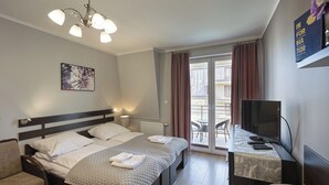 A hotel room with a bed, a desk with a TV, a chair, a window with curtains, and a painting on the wall. - Pegaz Apartments by Baltic Home (Swinoujscie)