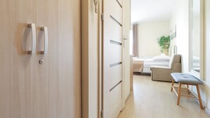 A hotel room with a wooden floor, a bed with a white headboard, a bedside table, a chair, and a window with curtains. - Pegaz Apartments by Baltic Home (Swinoujscie)