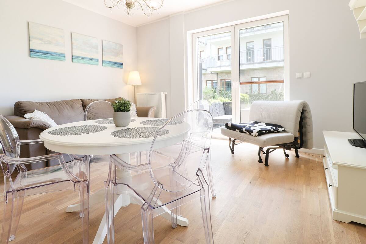 Ein modernes Wohnzimmer mit einem runden Glastisch, klaren Stühlen, einer Couch und einem Fernseher.