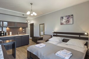 Una habitación de hotel moderna con kitchenette, una cama y toallas ordenadas, una silla y un cuadro floral grande en la pared.