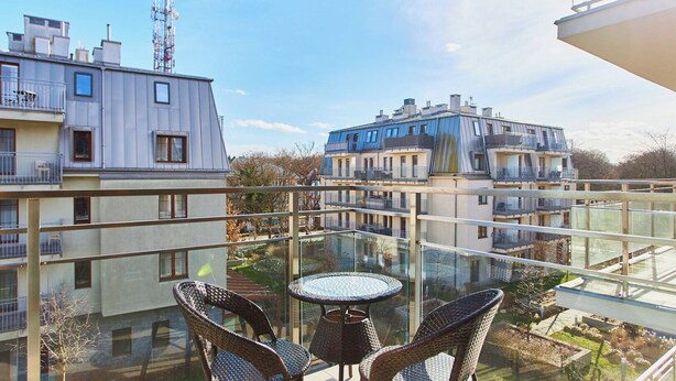 A glass-enclosed balcony with wicker chairs and a small table, overlooking a residential area with modern buildings and a clear sky. - Pegaz Apartments by Baltic Home (Swinoujscie)