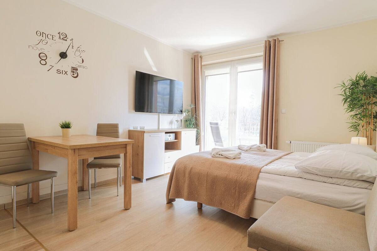Ein modernes Hotelzimmer mit Schreibtisch aus Holz, einem Bett mit beiger Decke, einem an der Wand montierten Fernseher und einem großen Fenster mit Vorhängen.