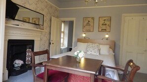 A bedroom with a bed, a wooden table, two chairs, a fireplace, and wall-mounted artwork. - Ryvington Apartments (Abergavenny)