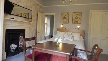 A bedroom with a bed, a wooden table, two chairs, a fireplace, and wall-mounted artwork.