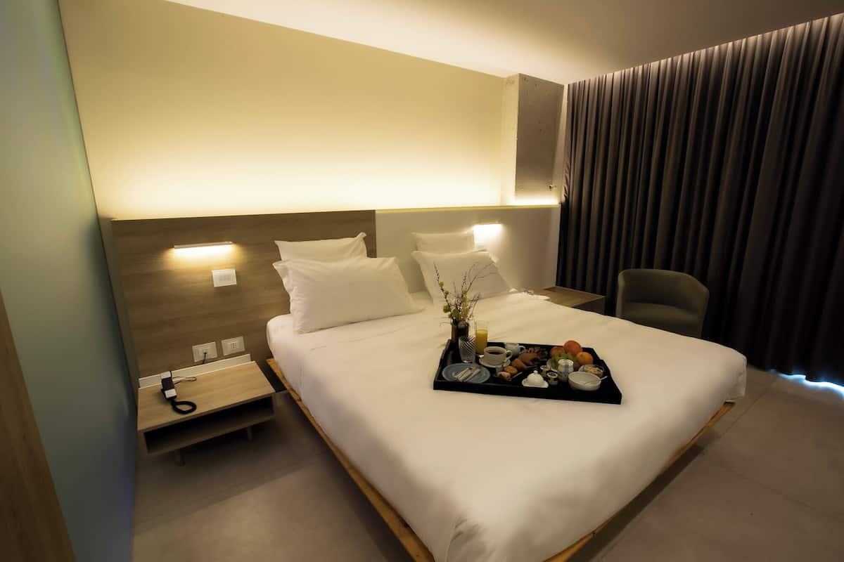 Ein Hotelzimmer mit einem ordentlich bezogenen Bett, einem Nachttisch mit Telefon und Ladegerät sowie einem Tablett mit Frühstücksgegenständen.