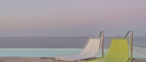 Two lounge chairs by a poolside with a view of the ocean.