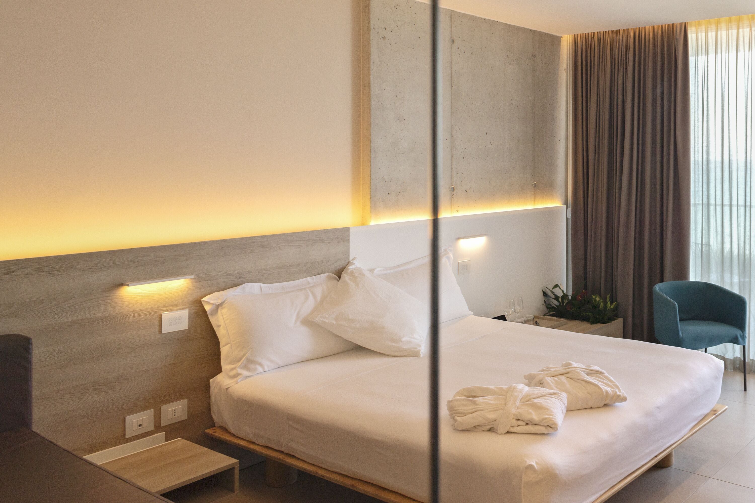 Une chambre d’hôtel moderne avec un grand lit, des tables de chevet, une petite table, une plante en pot et une vue sur l’extérieur par une fenêtre avec des rideaux.