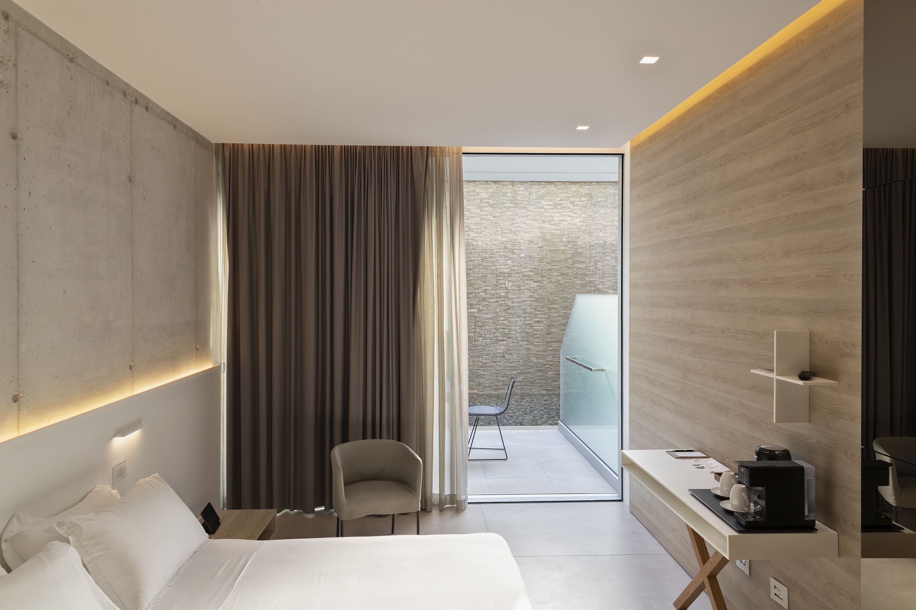 A modern hotel room with a bed, a desk with a coffee maker, a chair, and a view of a balcony.