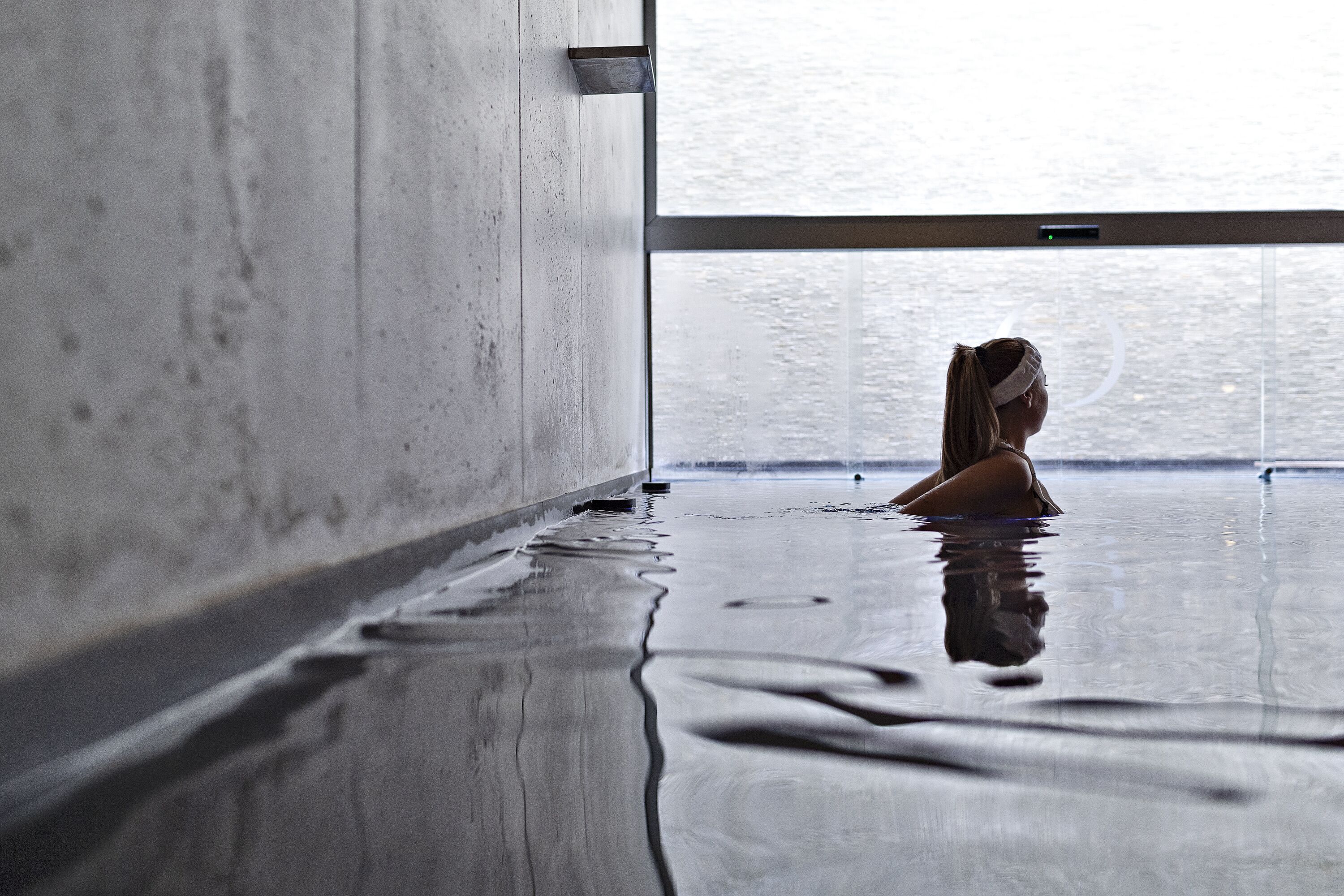 A person sitting in a wet indoor pool area with a concrete wall and a large window.