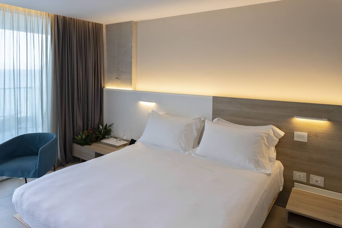 Ein modernes Hotelzimmer mit einem großen Bett, Nachttischen, einem Sessel und Blick ins Freie durch transparente Vorhänge.