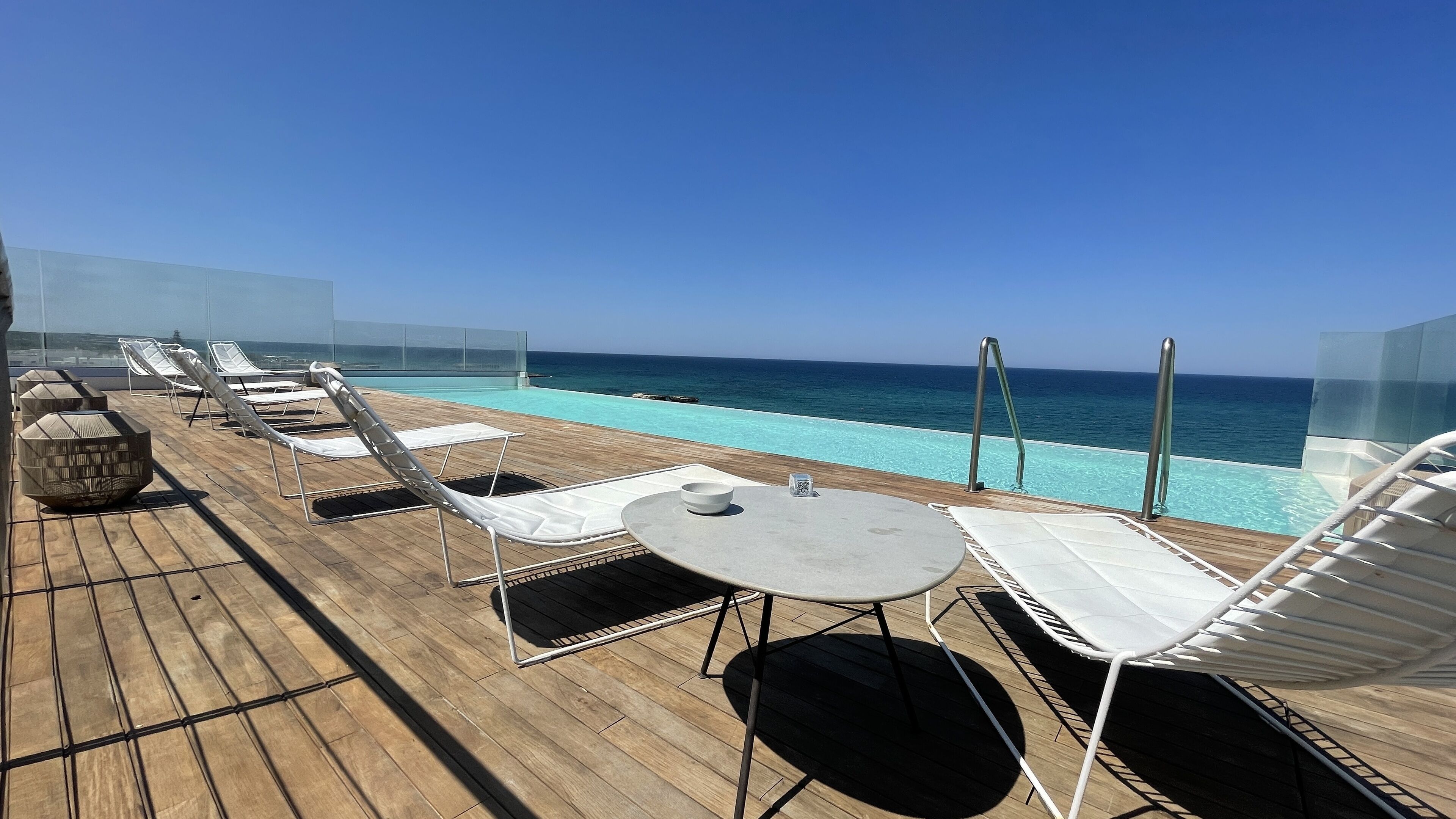 Terraço com espreguiçadeiras e mesa, com vista para uma piscina e o oceano.