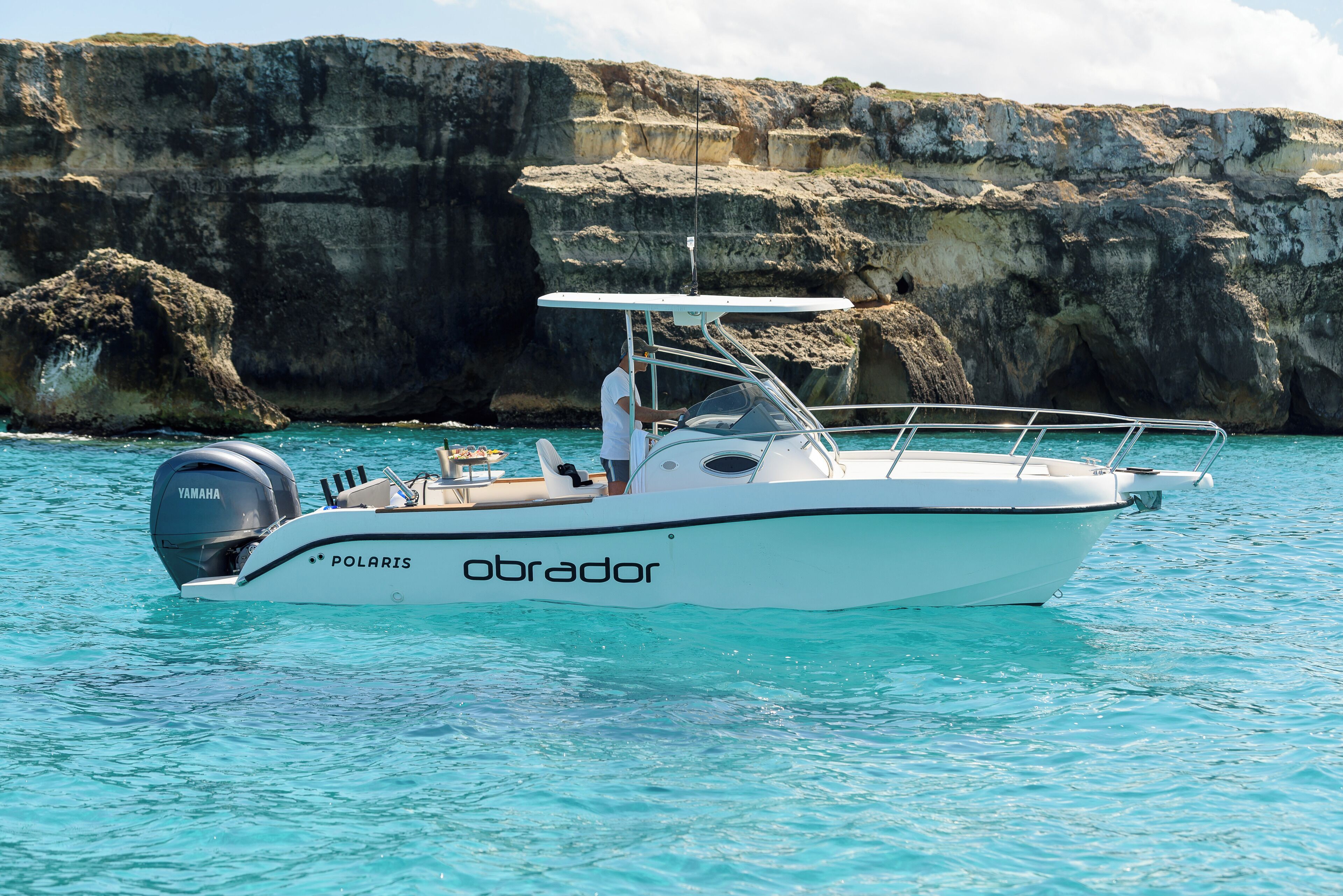 A small, white motorboat named 'Obrador' with a Yamaha outboard motor, floating on clear turquoise water near rocky cliffs.
