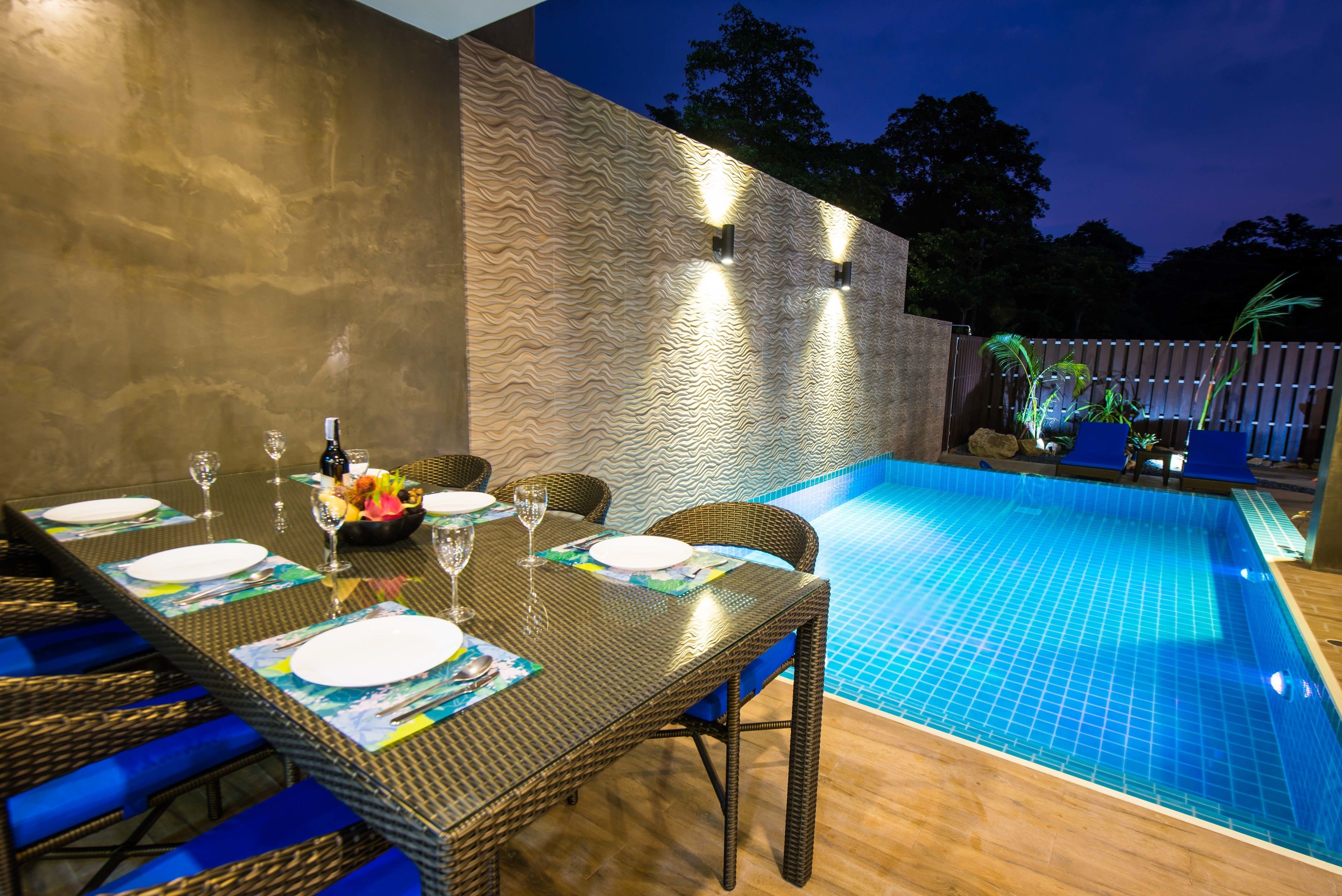 A dining area with a poolside table set for two, a pool with blue tiles, and a textured wall.