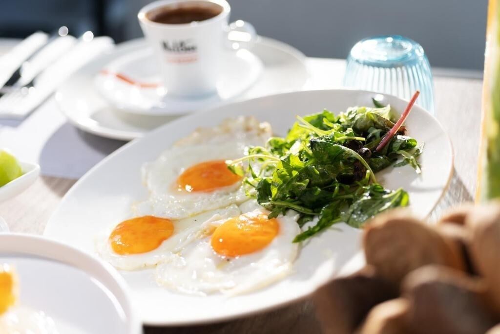 Ein Frühstücksteller mit zwei Spiegeleiern, einem Salat, einer Kaffeetasse und einem blauen Glas auf einem Tisch.