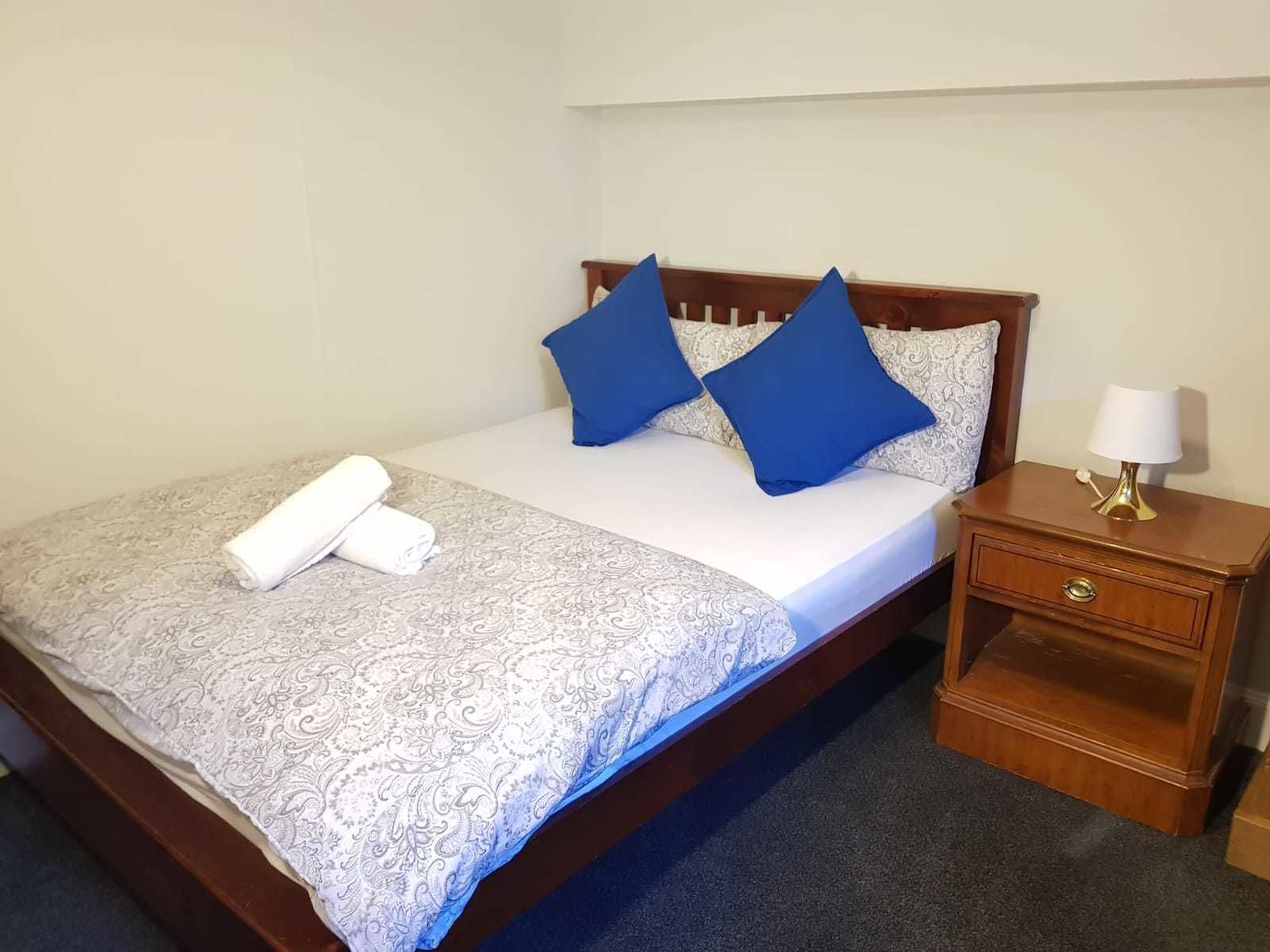 A neatly made bed with a patterned bedspread, two blue pillows, and a rolled white towel. There is a wooden nightstand with a lamp and a drawer.