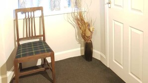 A corner room with a wooden chair, a window with patterned curtains, a vase with dried plants, and a white door.
