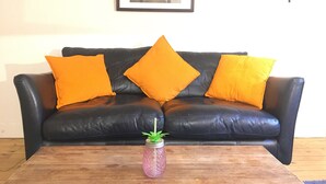A black leather sofa with three orange cushions, a framed picture on the wall, and a wooden coffee table with a pink vase.
