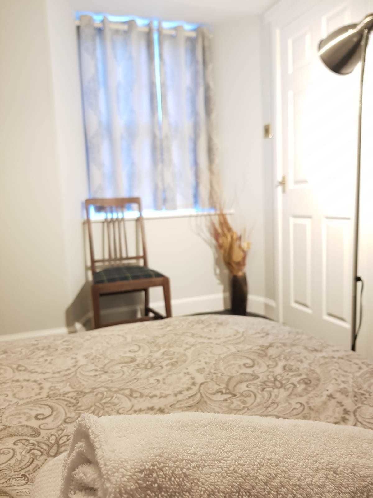 Ein Schlafzimmer mit einem gemusterten Bett, einem Sessel, einem Fenster mit Vorhängen und einer Stehlampe.