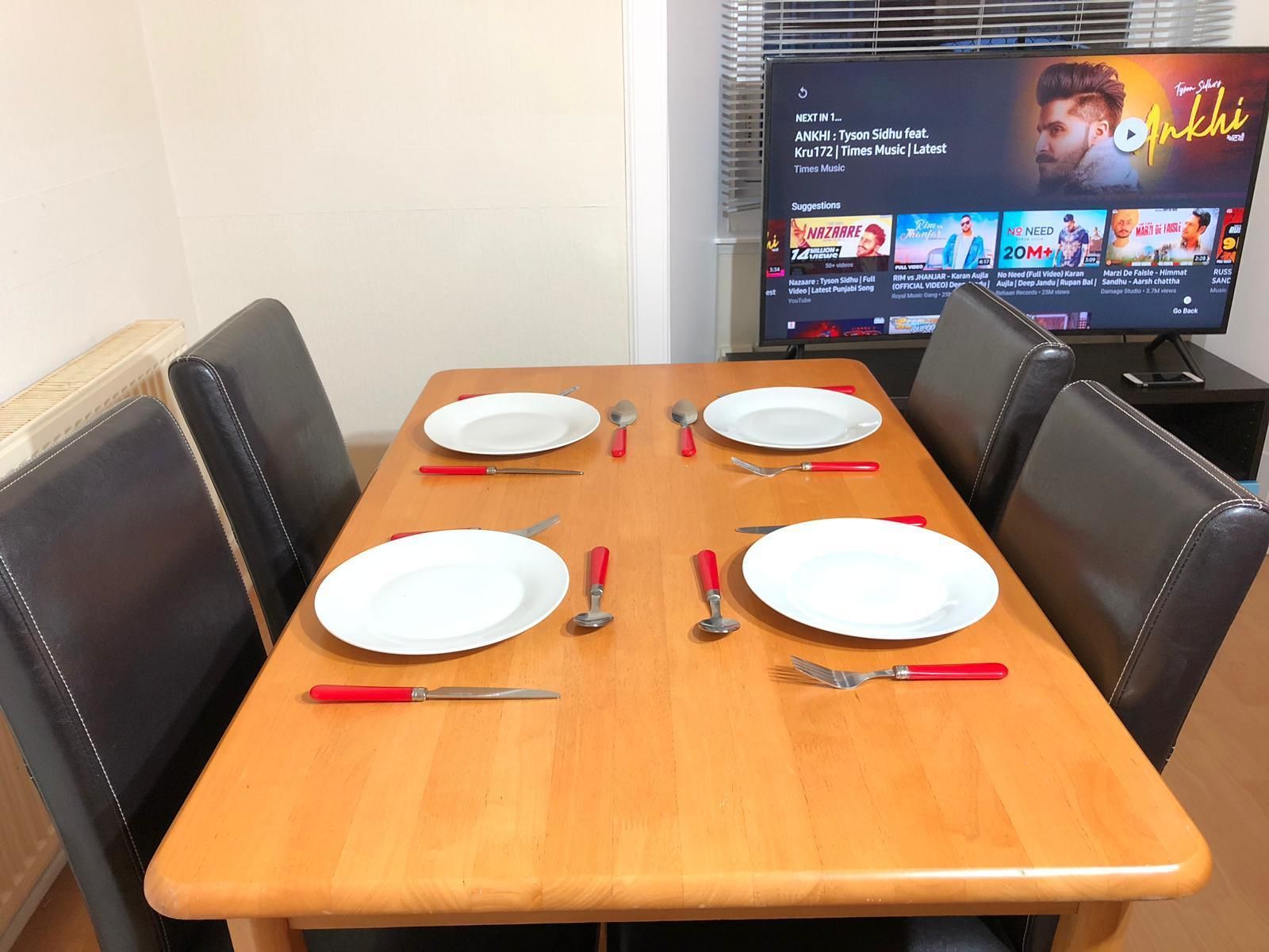 A dining table set with white plates, red-handled cutlery, and a television displaying a music video.