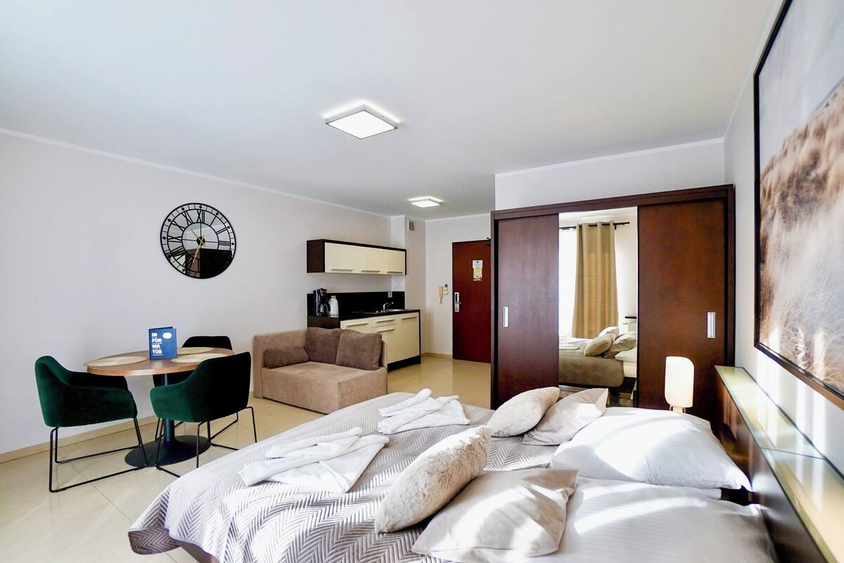 A hotel room with a bed, a desk with chairs, a sofa, a clock on the wall, and a door leading to another room.