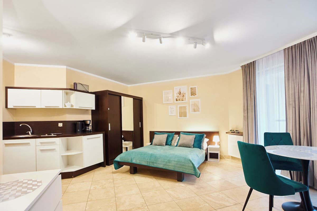 A modern hotel room with a bed, a desk, a chair, a dining table, and a kitchenette.