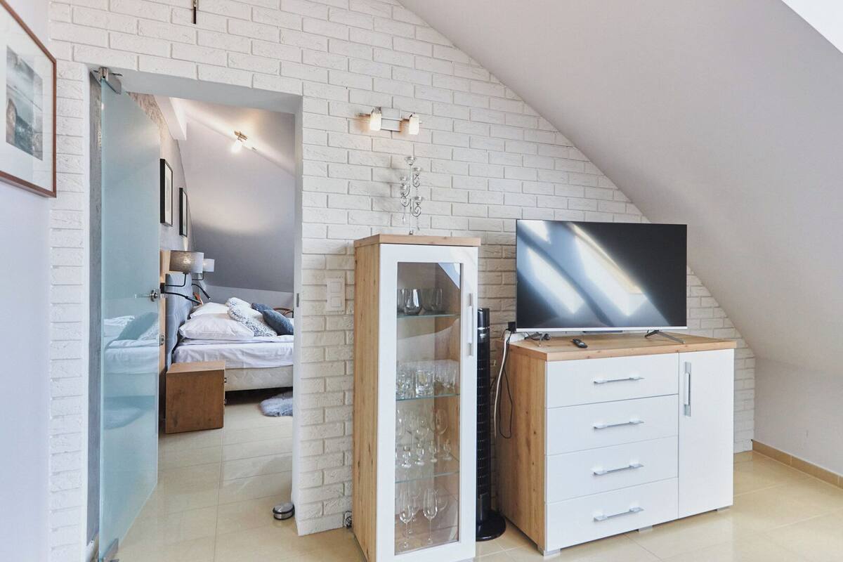 A modern bedroom with a sloped ceiling, a bed, a glass cabinet with wine glasses, a TV on a wooden cabinet, and a white brick wall.