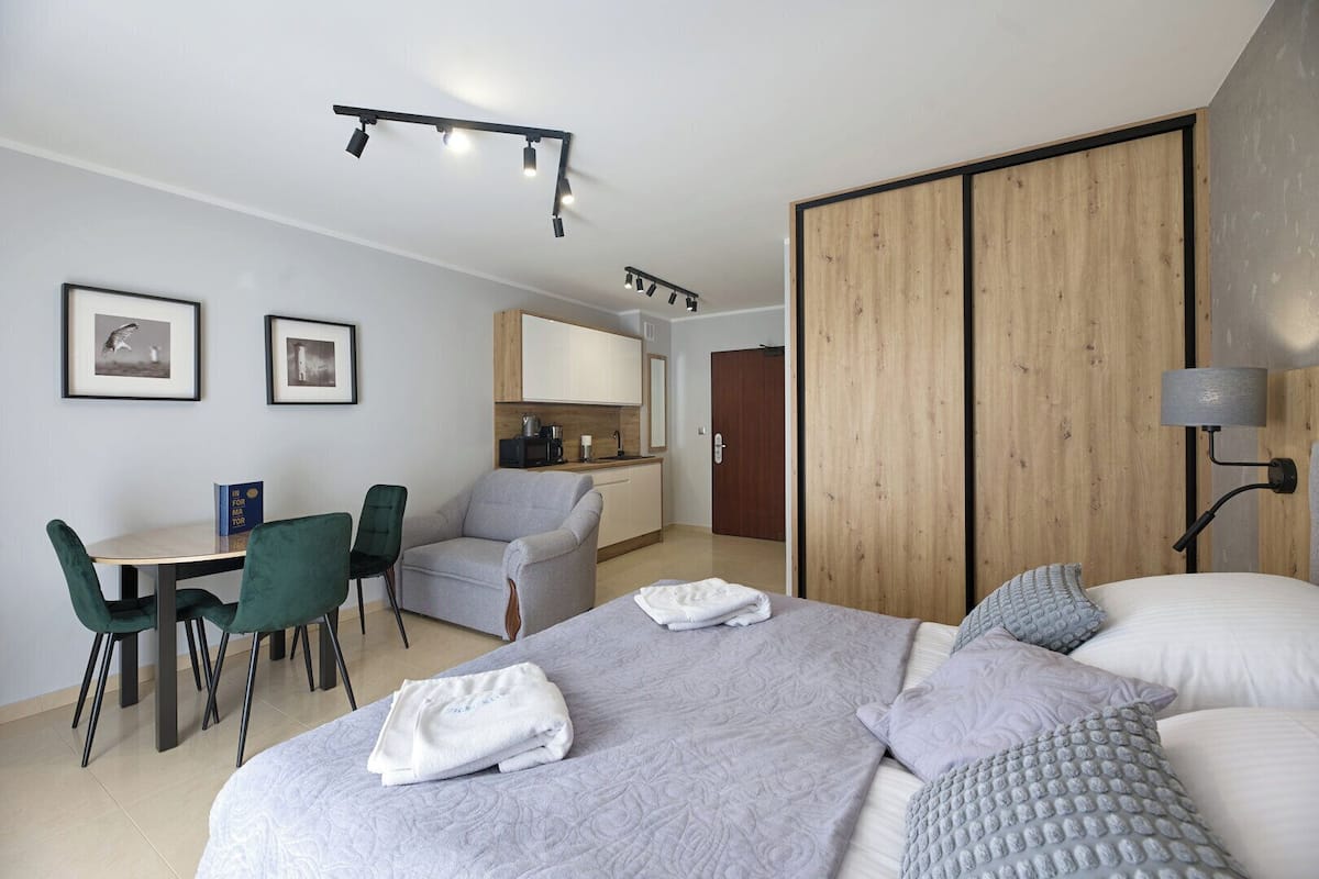 A modern hotel room with a large bed, a desk with a chair, a kitchenette, and a wardrobe.
