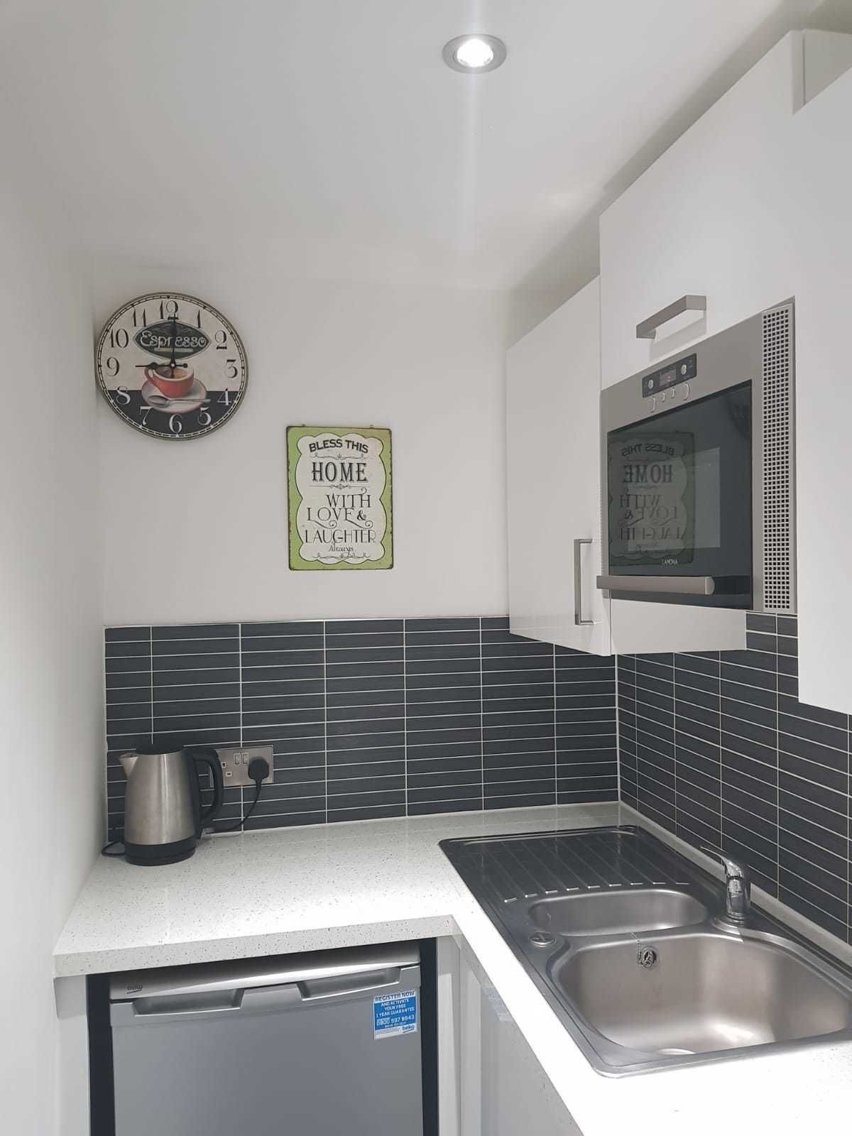A modern kitchen with a stainless steel sink, a kettle, and a microwave. There are two wall decorations, one of which is a clock.