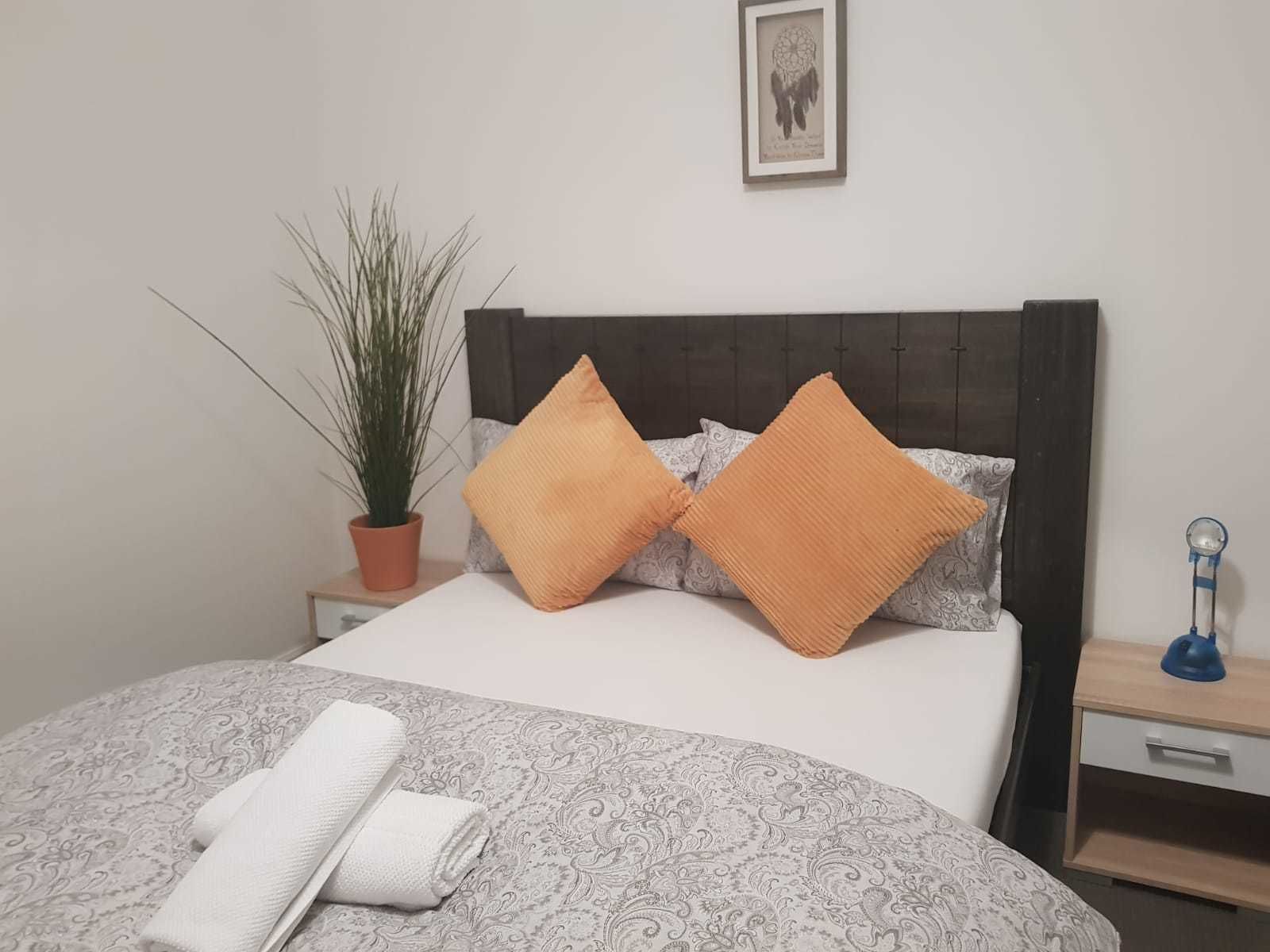A neatly made bed with patterned bedding, two orange pillows, a potted plant, and a bedside table with a lamp.