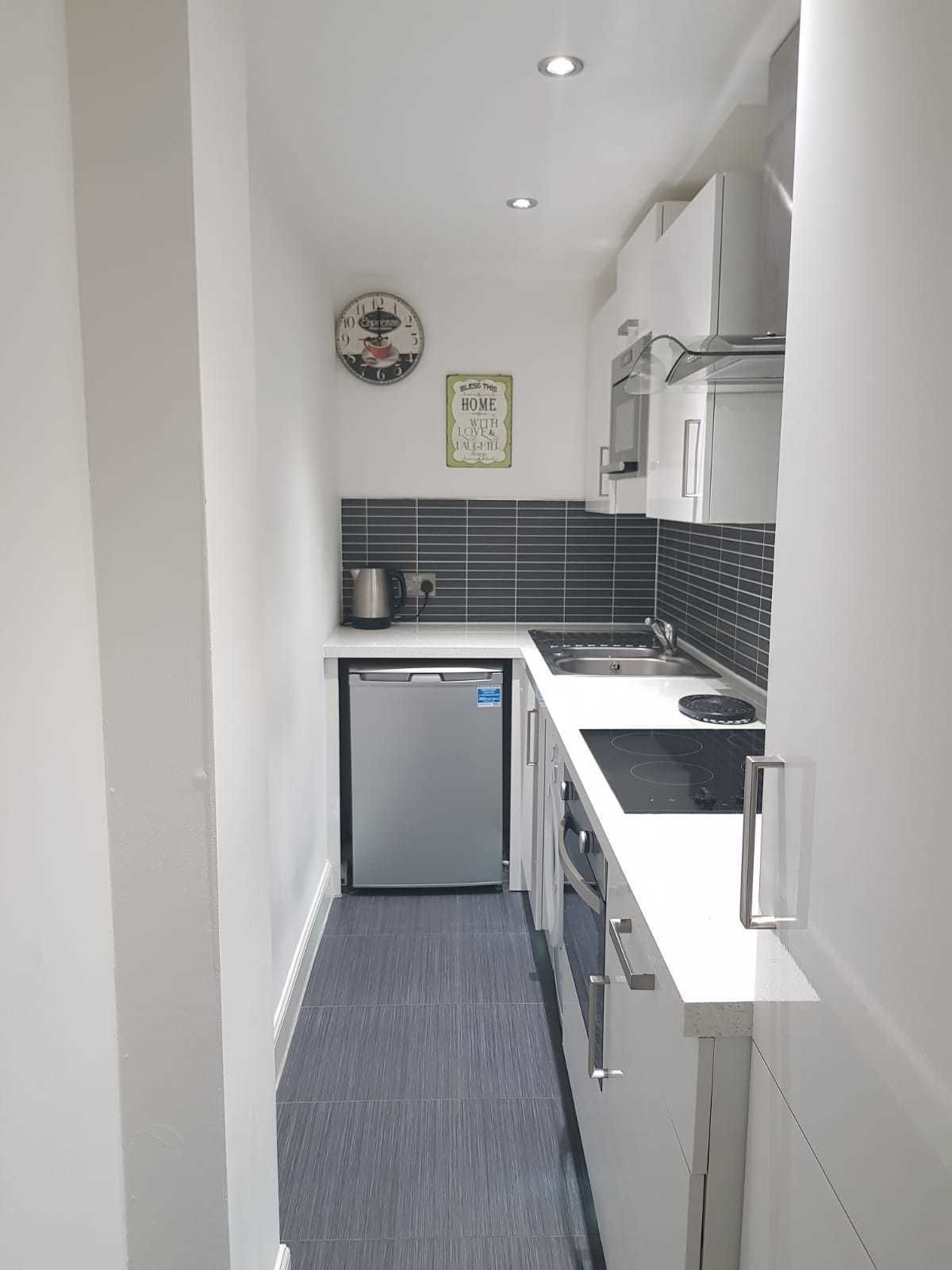 A compact kitchen with a refrigerator, stove, sink, and a clock on the wall.