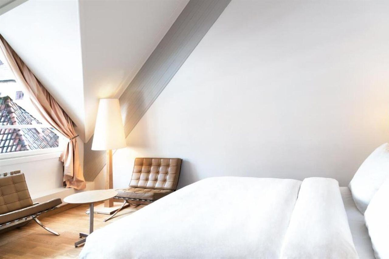 A hotel room with a bed, a chair, a small table, a lamp, and a view of buildings through a window.