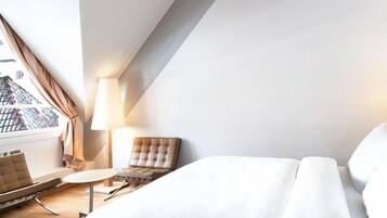 A hotel room with a bed, a chair, a small table, a lamp, and a view of buildings through a window.