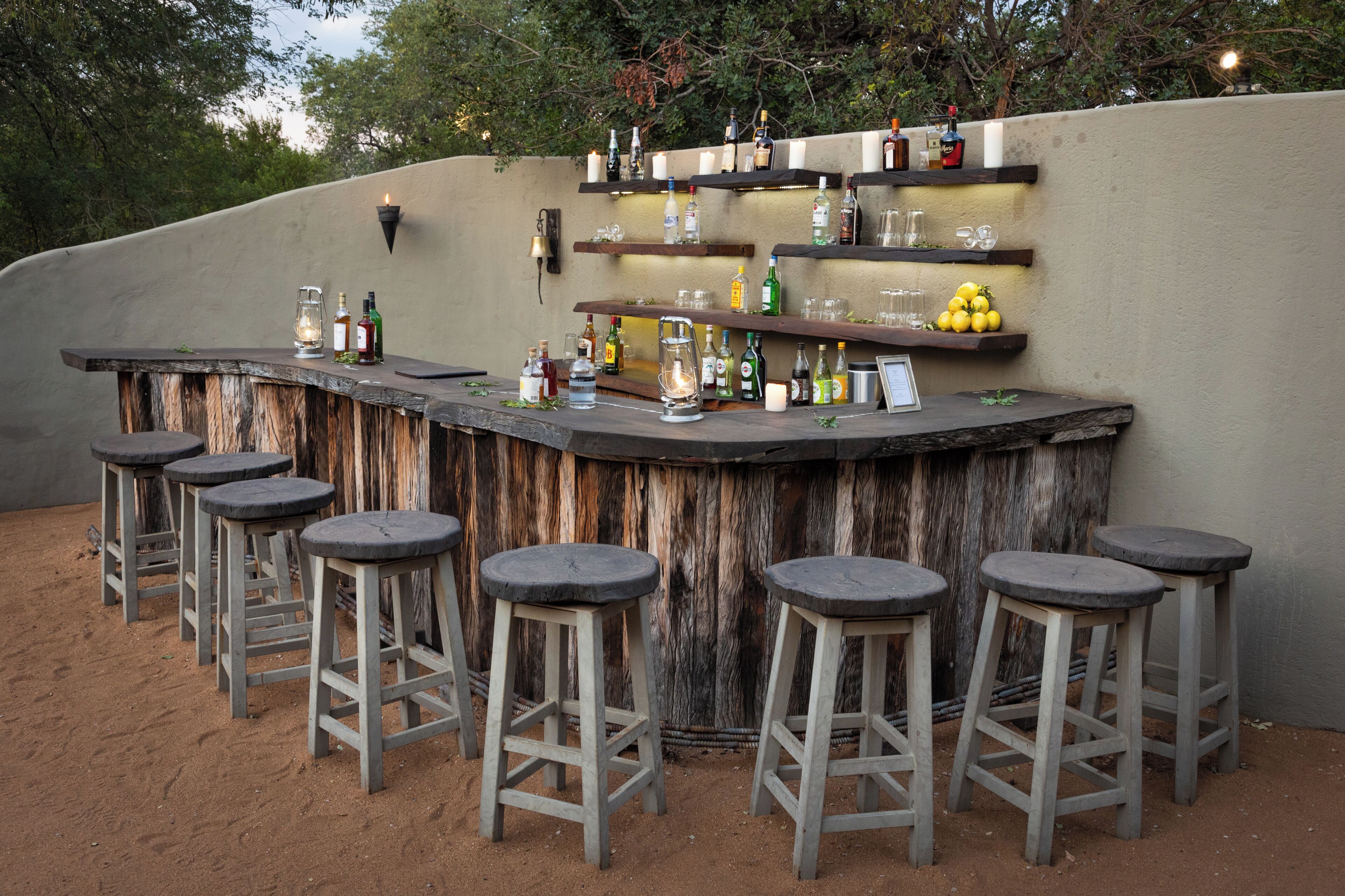 Un espace bar extérieur doté d’un comptoir en bois rustique, de plusieurs tabourets et d’étagères où sont disposées des bouteilles et des verres.